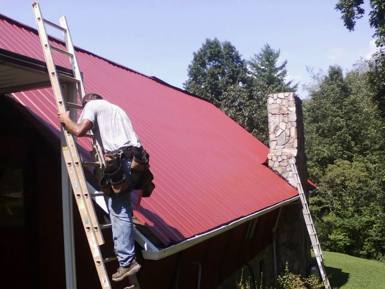 Roofer on a ladder beside a red metal roof and stone chimney — Soffit & Fascia Repair by Roofing Brothers in Risingsun
